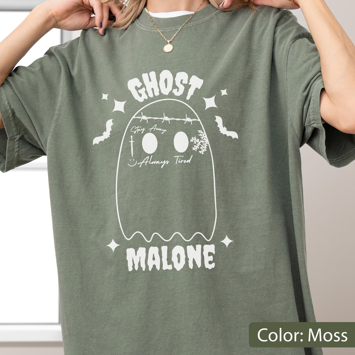 May include: Moss green t-shirt with a white ghost graphic. The ghost design includes the text "GHOST" above and "MALONE" below, with additional text and decorative elements within the ghost outline. The shirt has short sleeves.