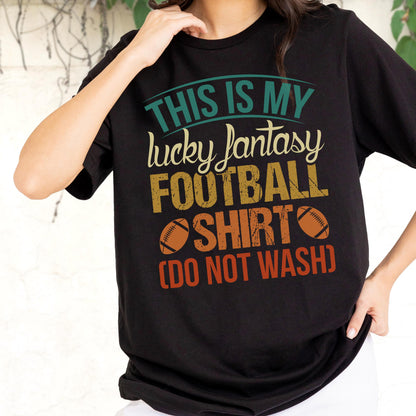 May include: A black short-sleeved t-shirt featuring the text "THIS IS MY lucky fantasy FOOTBALL SHIRT (DO NOT WASH)" in teal, cream, yellow, and orange, with two brown football illustrations flanking the word "SHIRT" on the front of the garment.