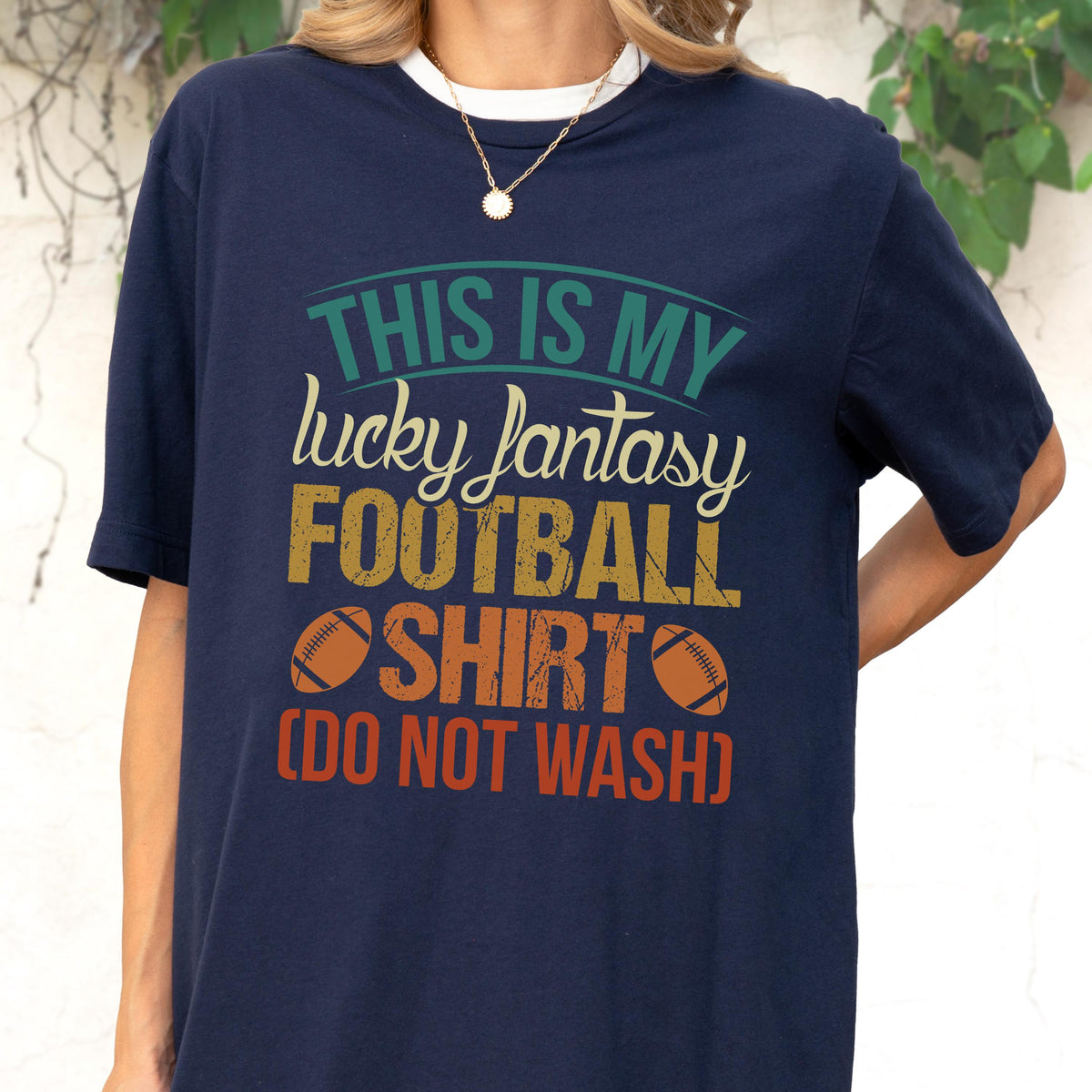 May include: A navy blue short-sleeve t-shirt featuring a graphic print with the text THIS IS MY lucky fantasy FOOTBALL SHIRT (DO NOT WASH) in teal, cream, and orange, accompanied by two brown football illustrations on the front of the garment.