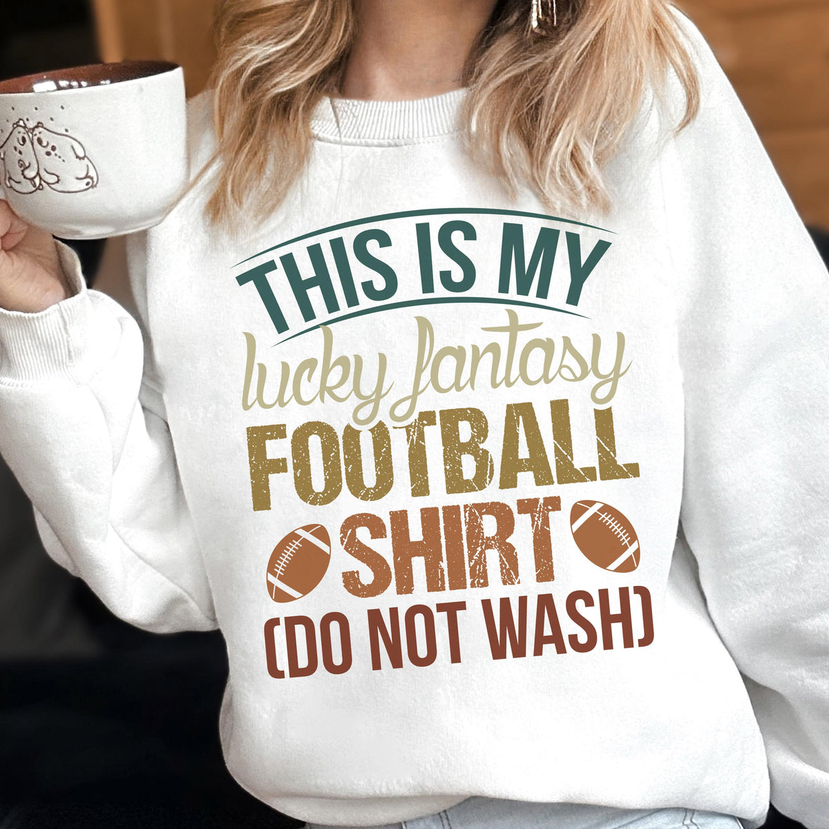 May include: White sweatshirt with the text "THIS IS MY lucky fantasy FOOTBALL SHIRT (DO NOT WASH)". The text is in green, tan, and brown colors, with two football graphics. A mug with a cartoon design is held in the image.