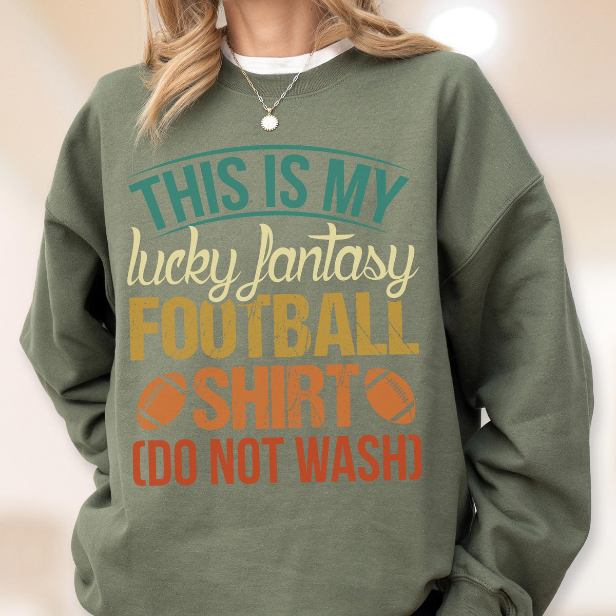 May include: Olive green sweatshirt with the text "THIS IS MY lucky fantasy FOOTBALL SHIRT (DO NOT WASH)". The text is in teal, yellow, orange, and white. Two orange footballs are printed on the shirt.