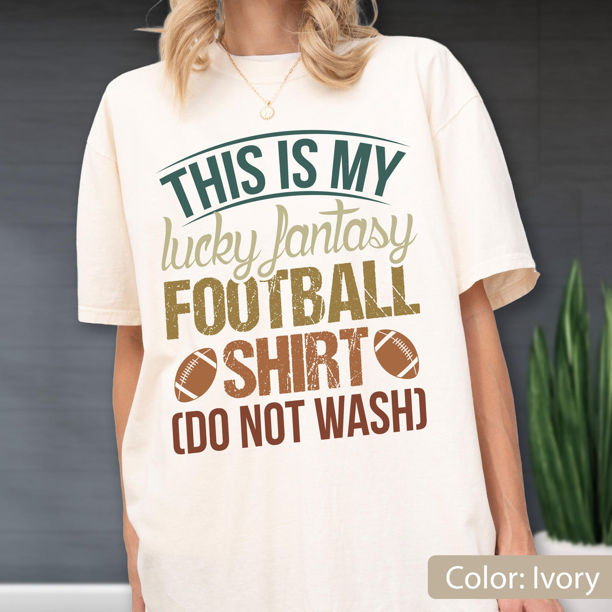 May include: An ivory short-sleeve t-shirt featuring the text "THIS IS MY lucky fantasy FOOTBALL SHIRT (DO NOT WASH)" in dark green, tan, and brown. Two brown football graphics are positioned on either side of the word "SHIRT" on the front.