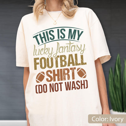 May include: An ivory short-sleeve t-shirt featuring the text "THIS IS MY lucky fantasy FOOTBALL SHIRT (DO NOT WASH)" in dark green, tan, and brown. Two brown football graphics are positioned on either side of the word "SHIRT" on the front.