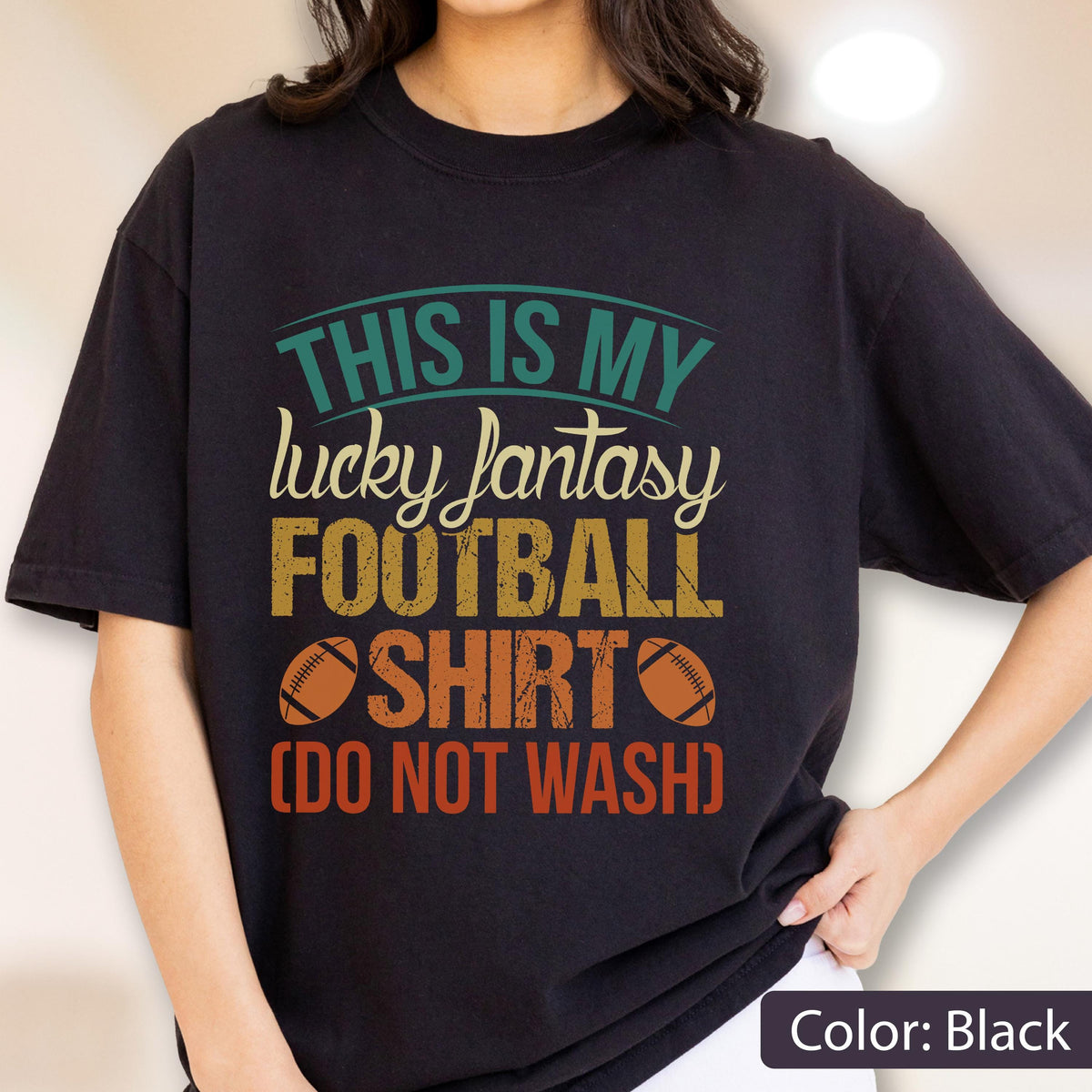 May include: A black short-sleeve t-shirt featuring a graphic print with the text "THIS IS MY lucky fantasy FOOTBALL SHIRT (DO NOT WASH)" in teal, cream, yellow, and orange, accompanied by two brown football illustrations on the front.