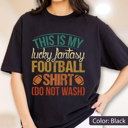 May include: A black short-sleeve t-shirt featuring a graphic print with the text "THIS IS MY lucky fantasy FOOTBALL SHIRT (DO NOT WASH)" in teal, cream, yellow, and orange, accompanied by two brown football illustrations on the front.