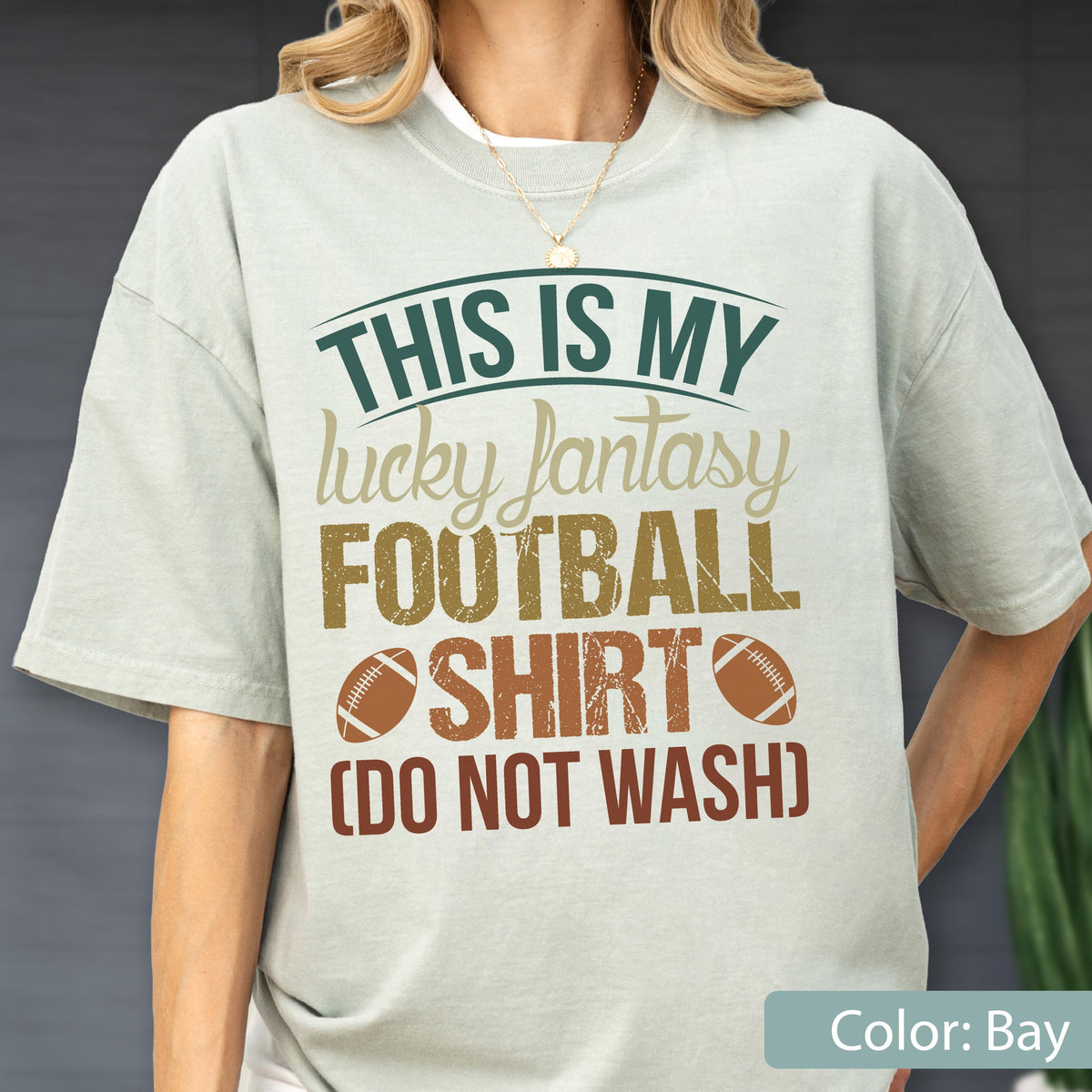 May include: A bay-colored short-sleeve t-shirt featuring the text THIS IS MY lucky fantasy FOOTBALL SHIRT (DO NOT WASH) in dark green, tan, and brown, accompanied by two brown football graphics on the front of the garment.