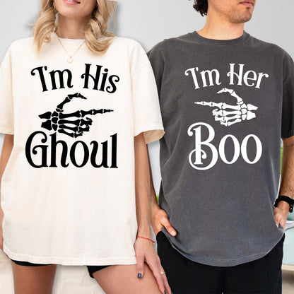 May include: A cream t-shirt with black text reading I'm His GHOUL and a skeleton hand graphic, paired with a charcoal gray t-shirt with white text reading I'm Her Boo and a skeleton hand graphic. Both shirts feature short sleeves and crew necks.