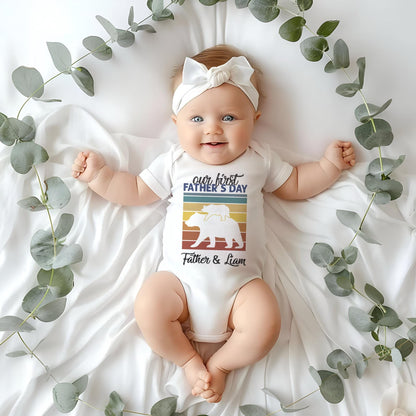 May include: A white baby onesie with a retro-style graphic of a bear and a cub. The text on the onesie reads 'our first father's day' and 'Father & Liam'.
