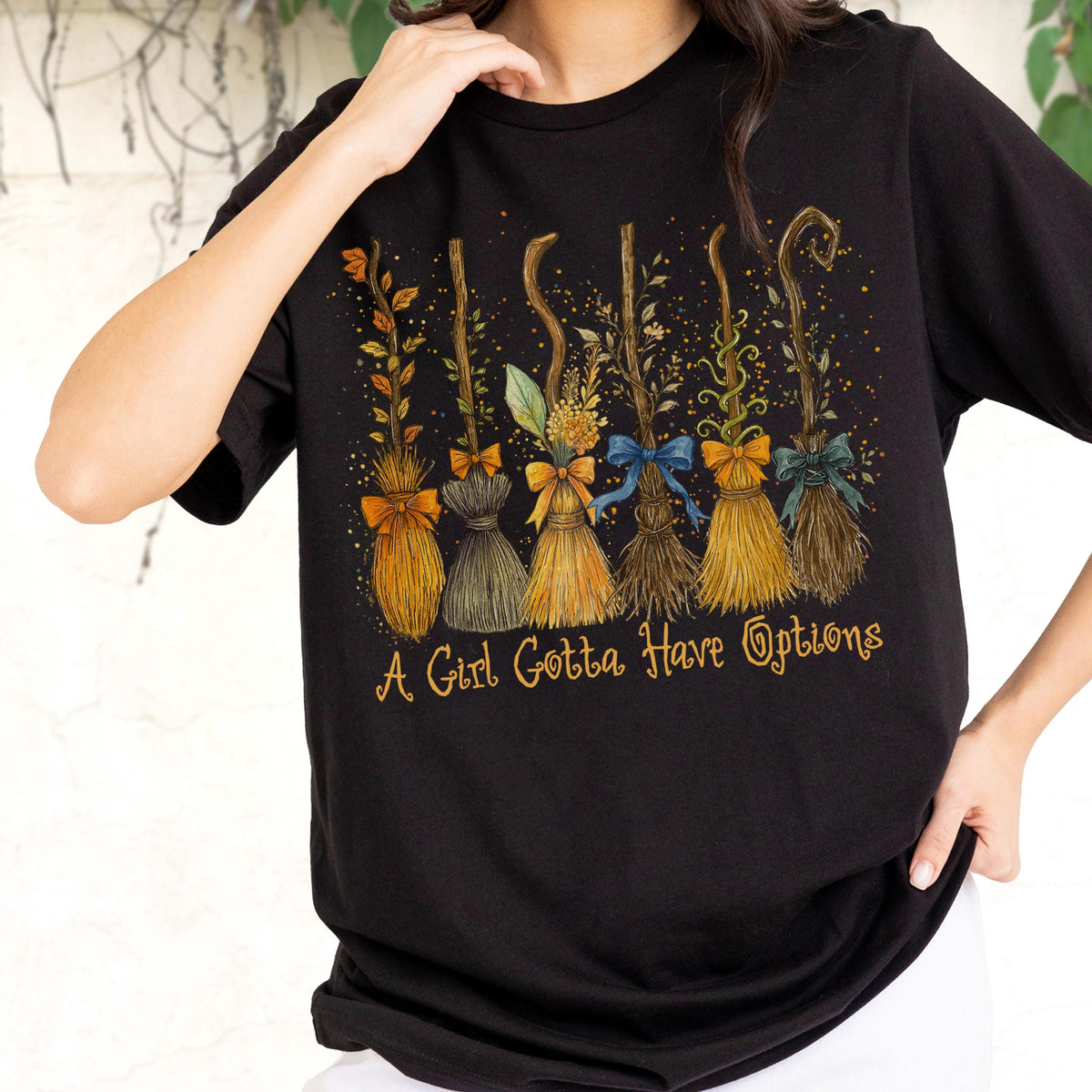 May include: A black short-sleeved t-shirt featuring a graphic of six distinct broomsticks decorated with various bows and foliage. Below the brooms, the text A Girl Gotta Have Options is printed in a stylized yellow cursive font.