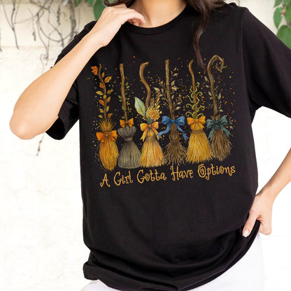 May include: A black short-sleeved t-shirt featuring a graphic of six distinct broomsticks decorated with various bows and foliage. Below the brooms, the text A Girl Gotta Have Options is printed in a stylized yellow cursive font.