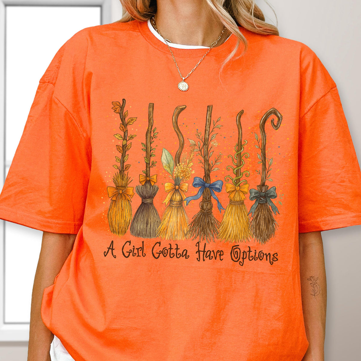 May include: An orange short-sleeved t-shirt featuring a graphic of six distinct broomsticks decorated with various bows and foliage. Below the broomsticks, the text "A Girl Gotta Have Options" is printed in a dark brown, stylized cursive font.