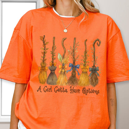 May include: An orange short-sleeved t-shirt featuring a graphic of six distinct broomsticks decorated with various bows and foliage. Below the broomsticks, the text "A Girl Gotta Have Options" is printed in a dark brown, stylized cursive font.