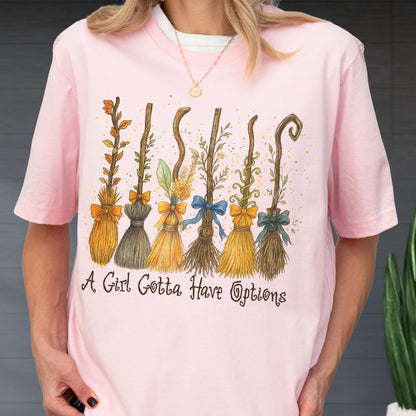 May include: A light pink short-sleeved t-shirt featuring a graphic of six distinct broomsticks decorated with various bows and foliage. Below the illustration, the text reads A Girl Gotta Have Options in a dark brown, cursive-style font.