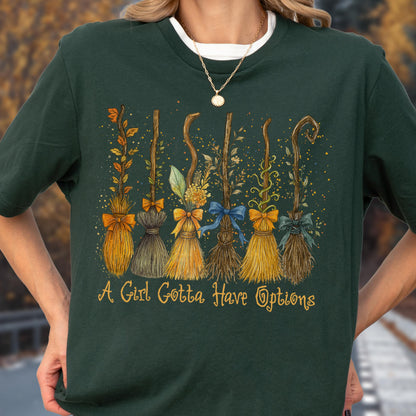 May include: A dark green t-shirt featuring a graphic of six distinct broomsticks decorated with various leaves, flowers, and bows. Below the brooms, the text "A Girl Gotta Have Options" is printed in a yellow, cursive-style font.