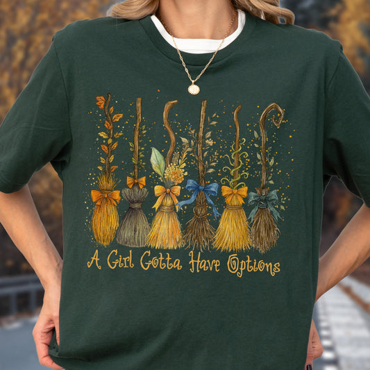 May include: A dark green t-shirt featuring a graphic of six distinct broomsticks decorated with various leaves, flowers, and bows. Below the brooms, the text "A Girl Gotta Have Options" is printed in a yellow, cursive-style font.