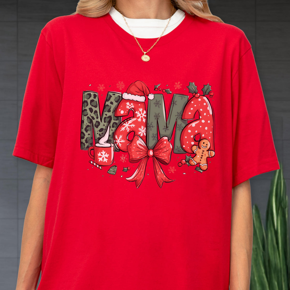 May include: Red t-shirt with the word "MAMA" in a festive Christmas design. The letters feature leopard print, snowflakes, and a Santa hat. A red bow, gingerbread man, and a cup of cocoa complete the holiday theme.