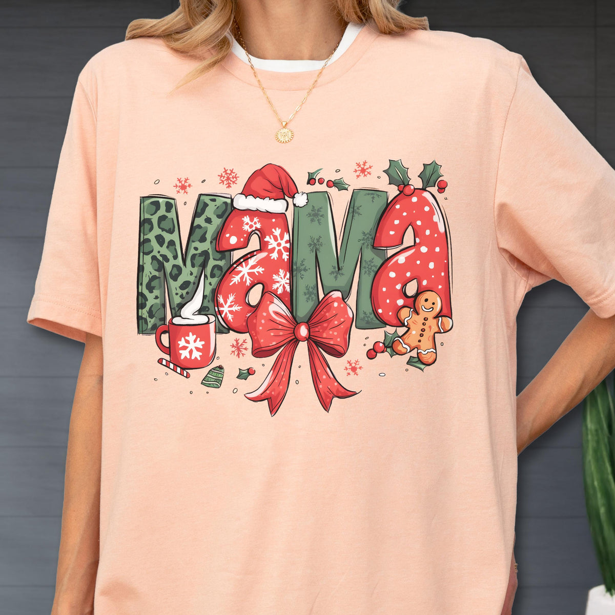 May include: Peach-colored t-shirt with the word "MAMA" in a Christmas-themed design. The letters feature leopard print, snowflakes, and red polka dots. A red bow, gingerbread man, and holly complete the festive look.