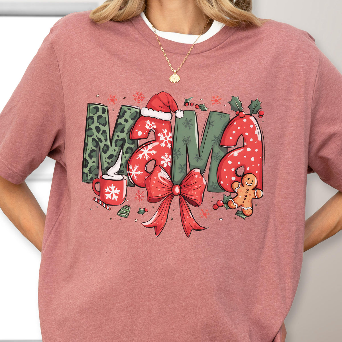 May include: A dusty rose-colored t-shirt featuring a festive "MAMA" design. The letters are decorated with Christmas elements: leopard print, snowflakes, a Santa hat, a red bow, and a gingerbread man. A mug of hot cocoa is also included.