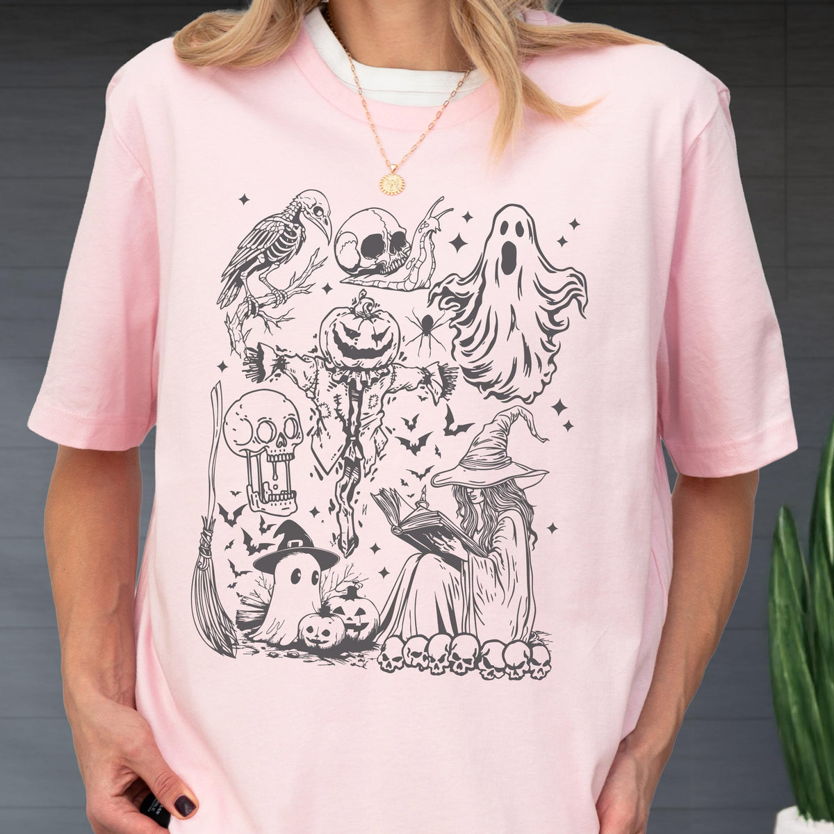 May include: A light pink t-shirt featuring a black and white Halloween-themed graphic. The design includes a crow skeleton, skulls, a ghost, a scarecrow, a witch reading a book, bats, and pumpkins. The shirt has short sleeves and a crew neck.