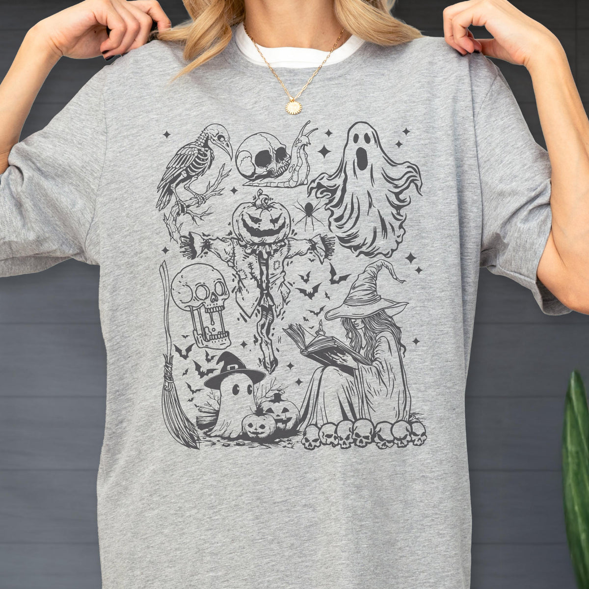 May include: A light gray t-shirt featuring a black and white Halloween-themed graphic. The graphic includes a scarecrow with a pumpkin head, a witch reading a book, ghosts, skulls, bats, and a broom.