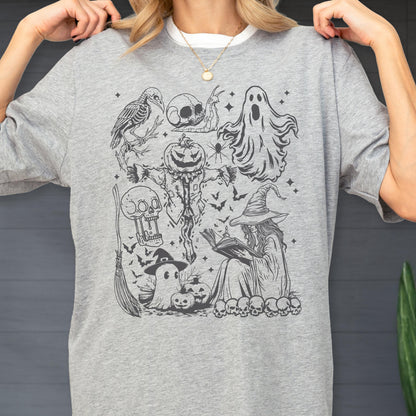 May include: A light gray t-shirt featuring a black and white Halloween-themed graphic. The graphic includes a scarecrow with a pumpkin head, a witch reading a book, ghosts, skulls, bats, and a broom.