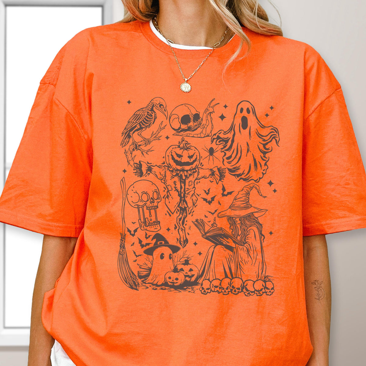 May include: Orange t-shirt with a black Halloween-themed graphic. The design includes a crow, skulls, a scarecrow, ghosts, bats, a witch reading a book, pumpkins, and a broom. The shirt is made of a soft material.
