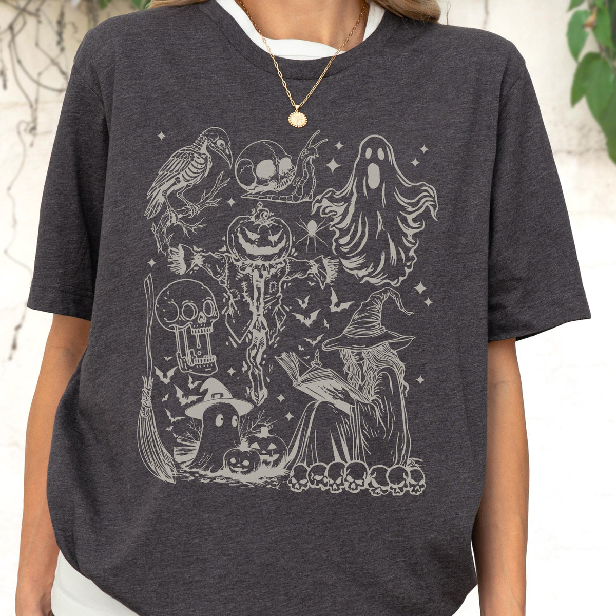 May include: Dark gray t-shirt featuring a detailed Halloween-themed graphic. The design includes a witch reading a book, ghosts, bats, skulls, a scarecrow, and a crow. The shirt has a crew neck and short sleeves, perfect for the spooky season.