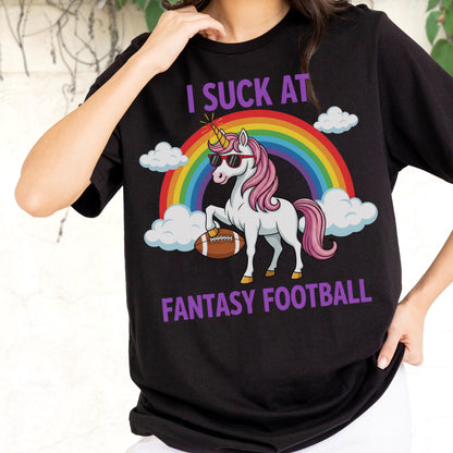 May include: A black t-shirt featuring a graphic of a white unicorn with a pink mane and tail, wearing sunglasses, holding a brown football, and standing in front of a rainbow and white clouds, with the purple text I SUCK AT FANTASY FOOTBALL.