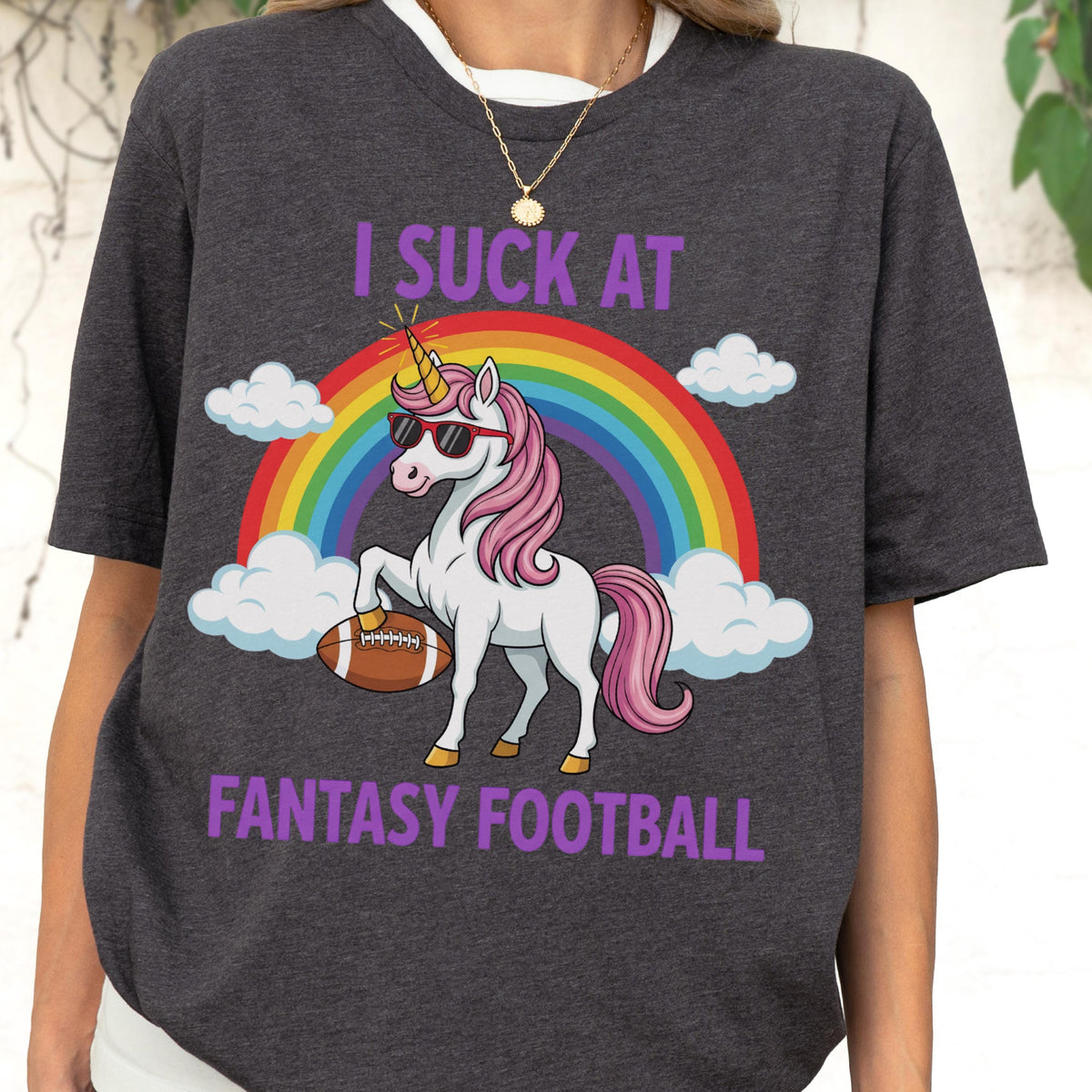 May include: A charcoal gray t-shirt featuring a graphic of a white unicorn with a pink mane and tail, wearing black sunglasses. The unicorn holds a brown football under one hoof. A rainbow and white clouds appear behind it with the text I SUCK AT FANTASY FOOTBALL.