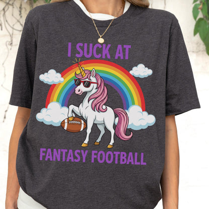 May include: A charcoal gray t-shirt featuring a graphic of a white unicorn with a pink mane and tail, wearing black sunglasses. The unicorn holds a brown football under one hoof. A rainbow and white clouds appear behind it with the text I SUCK AT FANTASY FOOTBALL.
