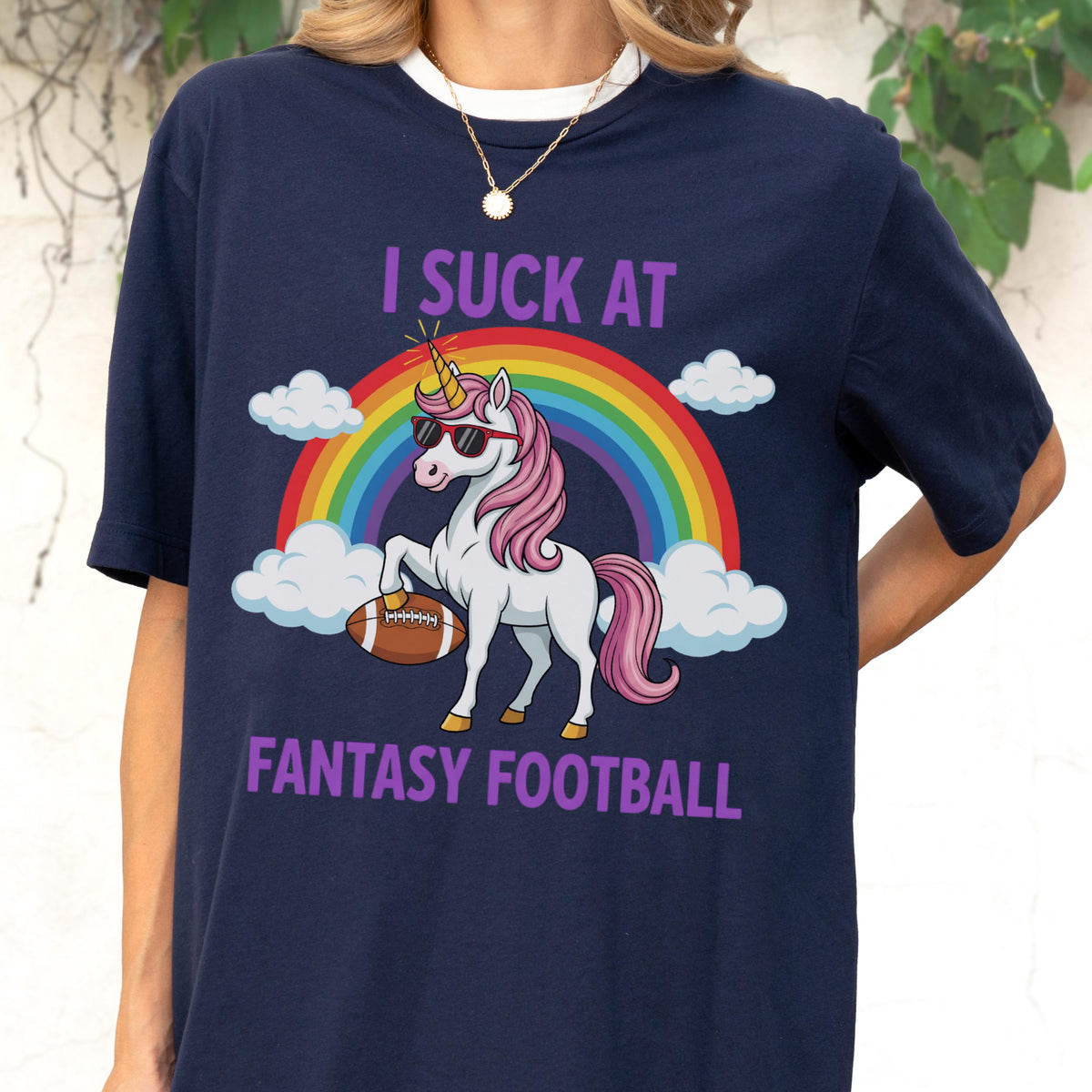 May include: A navy blue t-shirt featuring a graphic of a white unicorn with a pink mane and tail, wearing sunglasses and holding a brown football. Behind the unicorn is a colorful rainbow and white clouds. The text reads I SUCK AT FANTASY FOOTBALL.