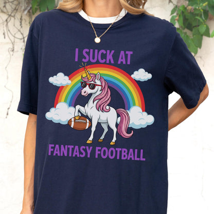 May include: A navy blue t-shirt featuring a graphic of a white unicorn with a pink mane and tail, wearing sunglasses and holding a brown football. Behind the unicorn is a colorful rainbow and white clouds. The text reads I SUCK AT FANTASY FOOTBALL.