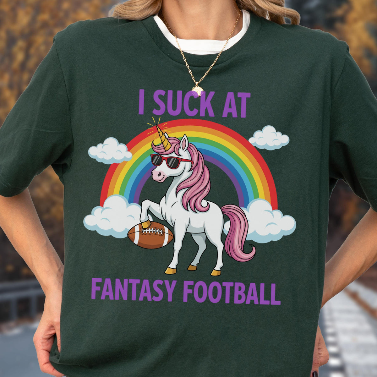 May include: A dark green short-sleeved t-shirt featuring a graphic of a white unicorn with a pink mane, wearing sunglasses and holding a brown football. Above and below the unicorn, the text reads I SUCK AT FANTASY FOOTBALL with a rainbow.