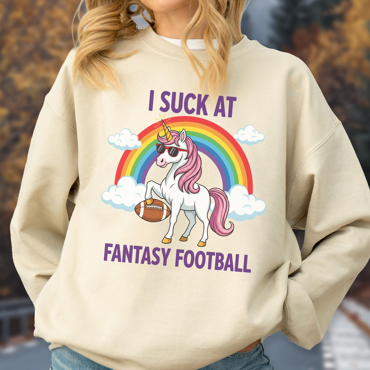 May include: A beige crewneck sweatshirt featuring a graphic of a white unicorn with a pink mane, wearing sunglasses and holding a brown football. Behind the unicorn is a colorful rainbow and white clouds. The text reads I SUCK AT FANTASY FOOTBALL.