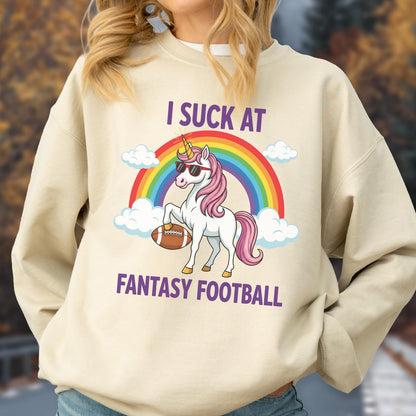 May include: A beige crewneck sweatshirt featuring a graphic of a white unicorn with a pink mane, wearing sunglasses and holding a brown football. Behind the unicorn is a colorful rainbow and white clouds. The text reads I SUCK AT FANTASY FOOTBALL.