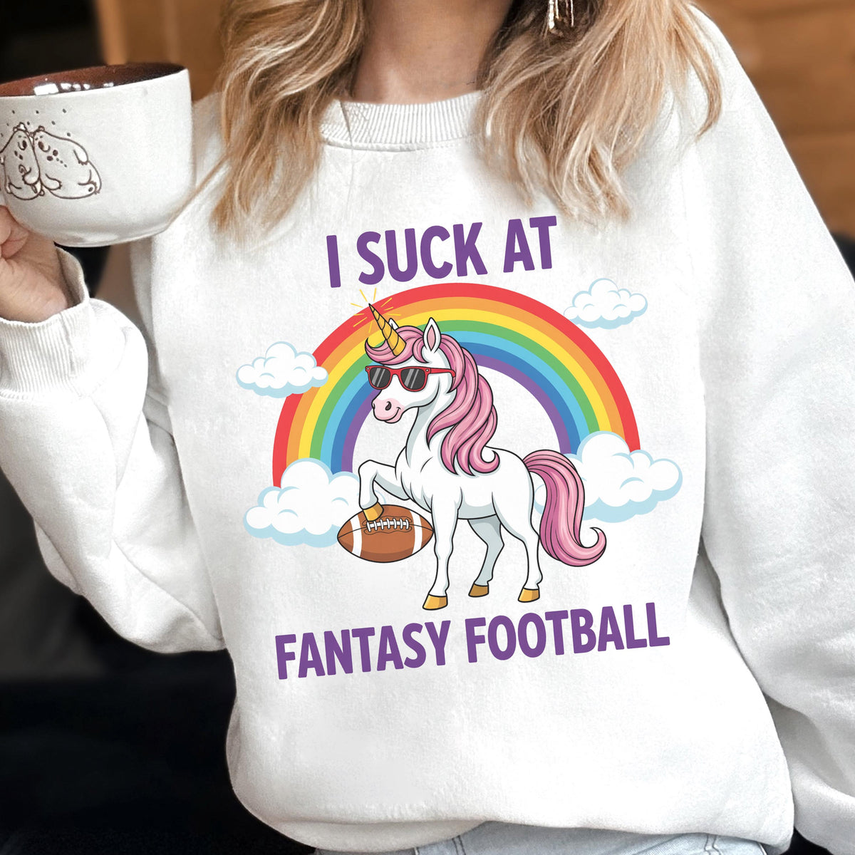 May include: A white crewneck sweatshirt featuring a graphic of a white unicorn with a pink mane and tail, wearing black sunglasses. The unicorn stands before a colorful rainbow and clouds, holding a brown football. Text reads I SUCK AT FANTASY FOOTBALL.