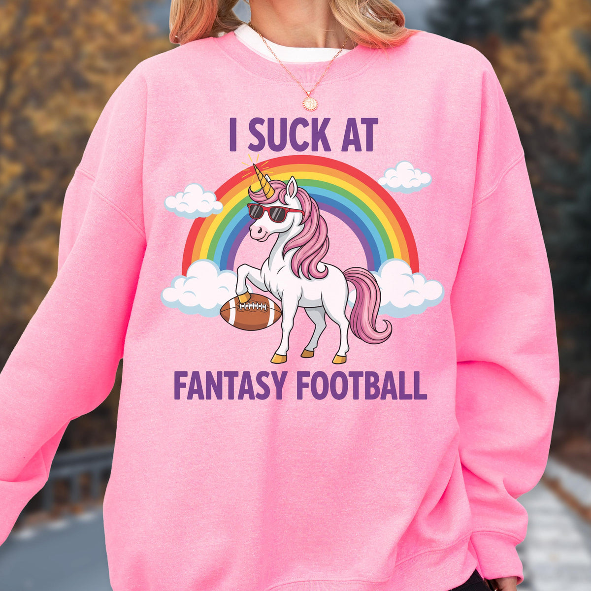 May include: A bright pink long-sleeve sweatshirt featuring a graphic of a white unicorn with a pink mane, wearing sunglasses and holding a brown football. Above and below the unicorn, the text reads I SUCK AT FANTASY FOOTBALL, with a rainbow and clouds.