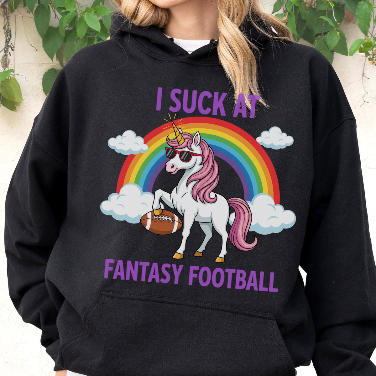 May include: A black hooded sweatshirt featuring a graphic of a white unicorn with a pink mane and tail, wearing sunglasses and holding a brown football. Behind the unicorn is a colorful rainbow and white clouds. The text reads I SUCK AT FANTASY FOOTBALL.