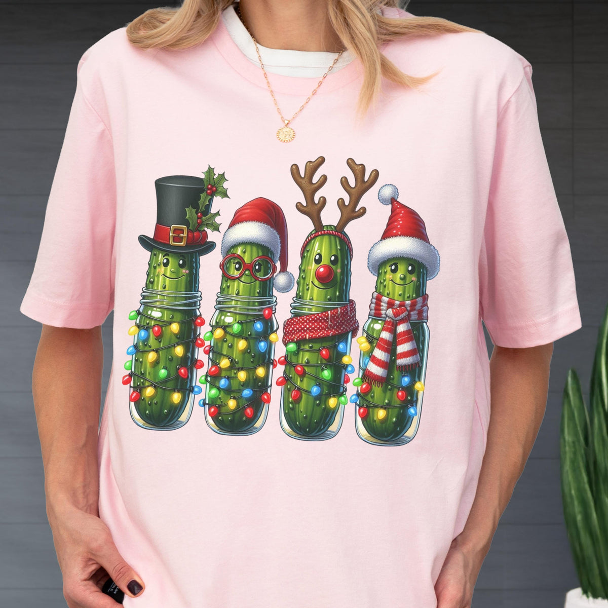 May include: Pink t-shirt featuring a whimsical illustration of four anthropomorphic pickles in glass jars, each adorned with festive holiday accessories. The pickles are wrapped in string lights and wear a top hat, Santa hats, reindeer antlers, and scarves.