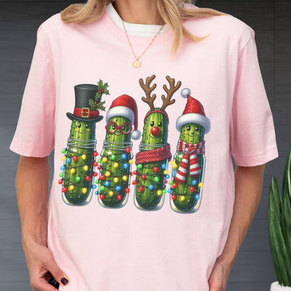 May include: Pink t-shirt featuring a whimsical illustration of four anthropomorphic pickles in glass jars, each adorned with festive holiday accessories. The pickles are wrapped in string lights and wear a top hat, Santa hats, reindeer antlers, and scarves.