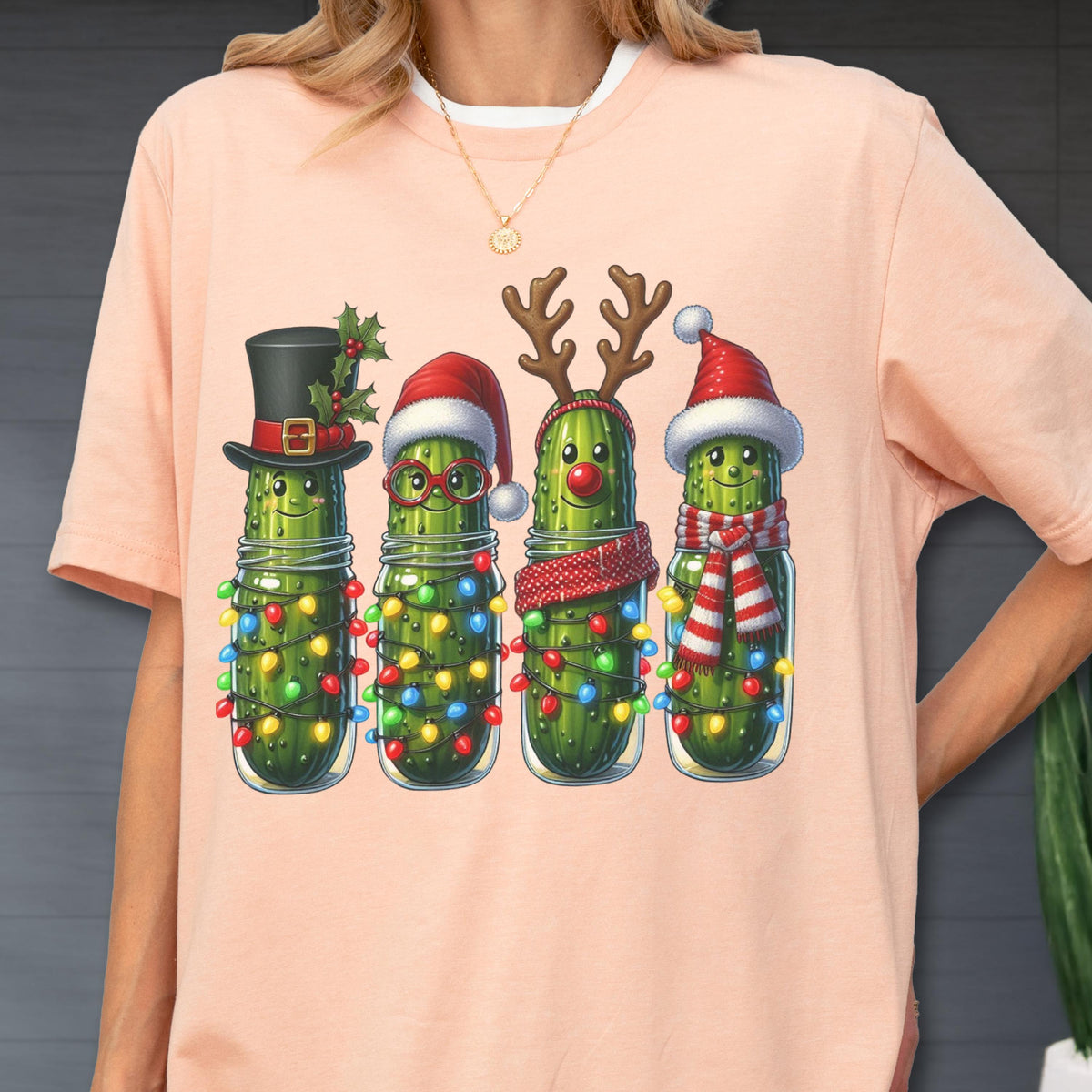 May include: A light pink t-shirt featuring a festive graphic of four anthropomorphic pickles in mason jars, each adorned with Christmas decorations. The pickles wear a top hat, Santa hats, reindeer antlers, and scarves, all surrounded by colorful string lights.