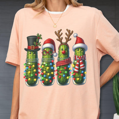 May include: A light pink t-shirt featuring a festive graphic of four anthropomorphic pickles in mason jars, each adorned with Christmas decorations. The pickles wear a top hat, Santa hats, reindeer antlers, and scarves, all surrounded by colorful string lights.