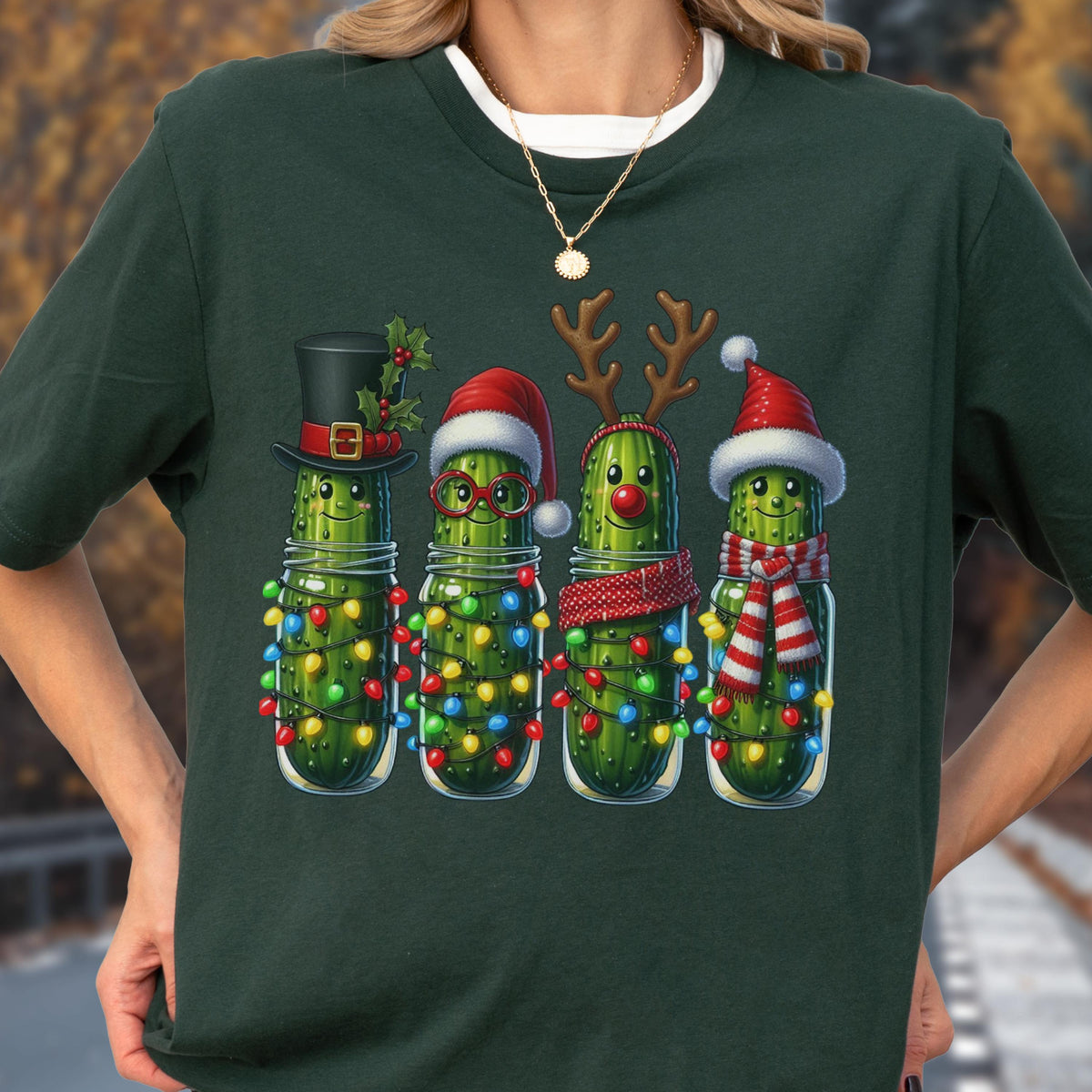May include: Dark green t-shirt featuring a festive design of four anthropomorphic pickles in glass jars, each adorned with Christmas decorations. The pickles wear a top hat, Santa hats, reindeer antlers, and a striped scarf, all illuminated by colorful string lights.
