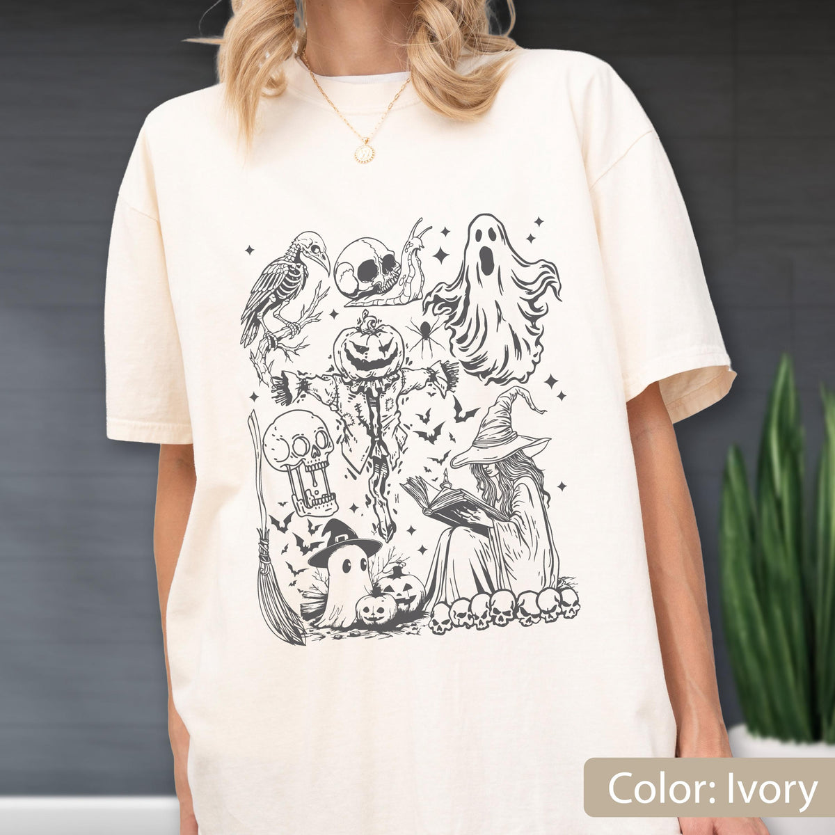 May include: Ivory-colored t-shirt featuring a black and white Halloween-themed graphic. The design includes a crow, skull, ghost, scarecrow, witch, bats, and pumpkins. The shirt is a relaxed fit.