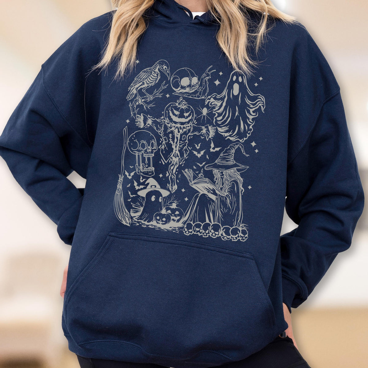 May include: Navy blue hoodie featuring a Halloween-themed graphic. The design includes a crow, skulls, a jack-o'-lantern, a ghost, a witch, bats, and other spooky elements. The hoodie has a drawstring hood and a front pocket.