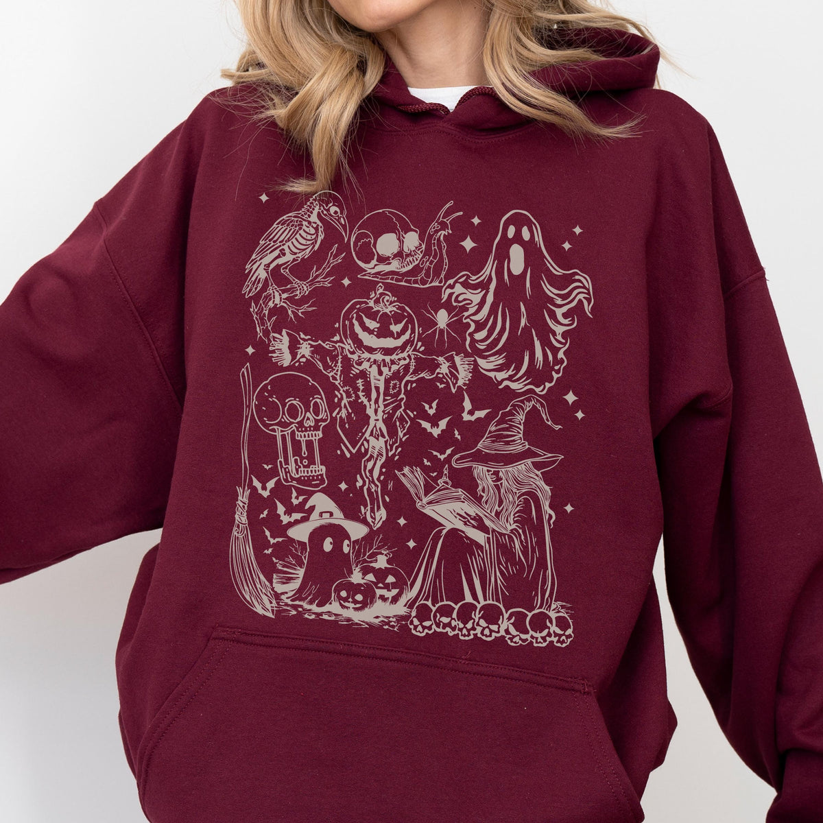May include: Burgundy hoodie featuring a detailed cream-colored Halloween-themed graphic. The design includes a witch, ghosts, pumpkins, bats, skulls, and a crow. The hoodie has a drawstring hood and a kangaroo pocket.