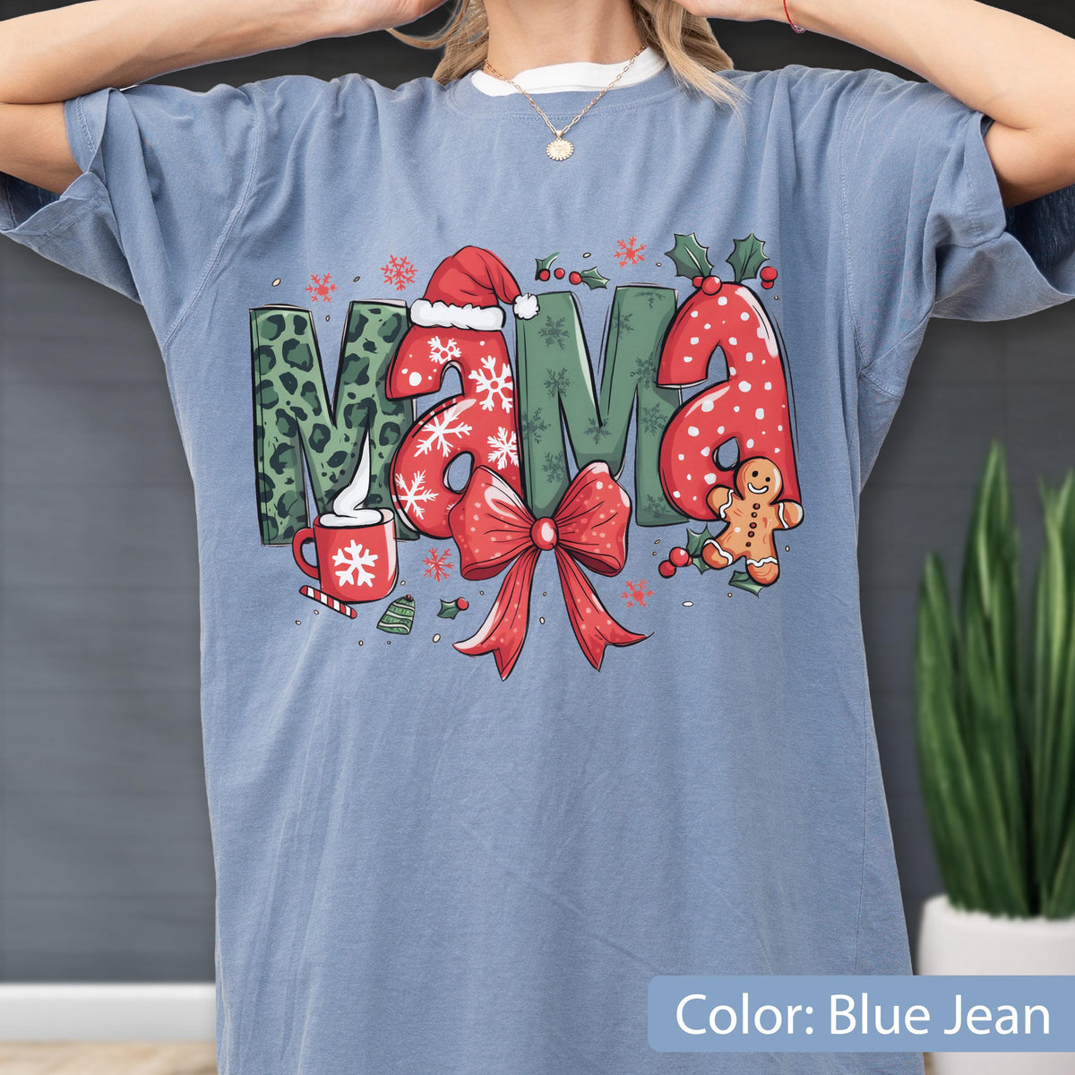 May include: Blue jean colored t-shirt with a Christmas-themed graphic. The word "MAMA" is displayed with festive designs: a Santa hat, snowflakes, a red bow, and a gingerbread man. The shirt also features a cup of hot cocoa.
