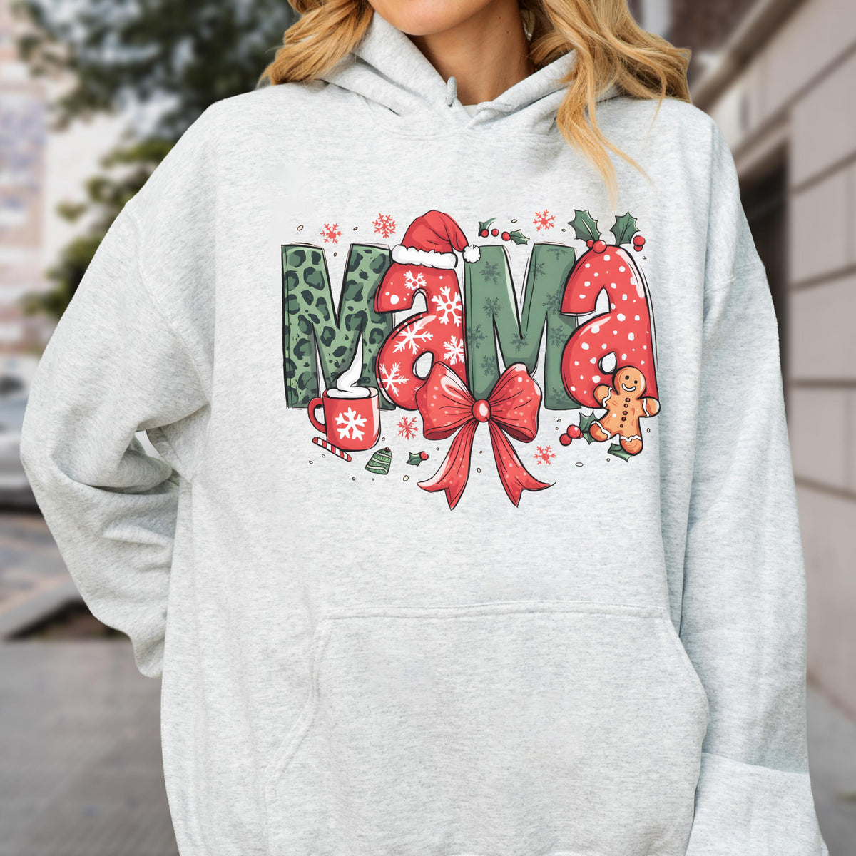 May include: A light gray hoodie with the word "MAMA" in a festive Christmas design. The letters feature a leopard print, snowflakes, and a Santa hat. A red bow, gingerbread man, and a mug of hot cocoa complete the holiday theme.