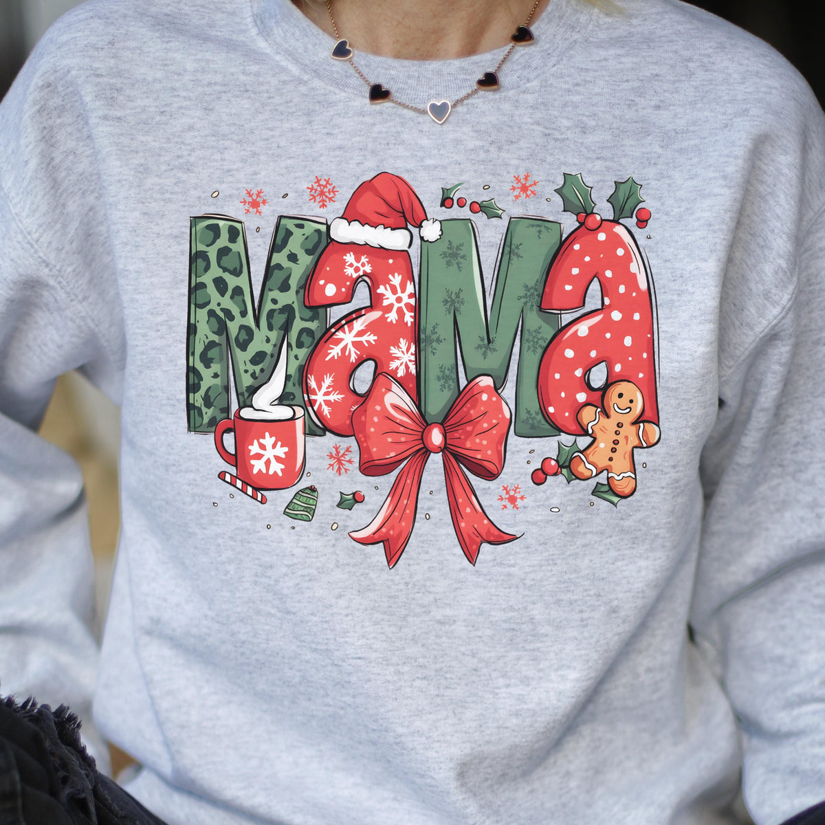 May include: A light gray sweatshirt with a festive "Mama" design. The letters feature a mix of leopard print, snowflakes, and red polka dots. A red bow, gingerbread man, and a mug of hot cocoa complete the holiday theme.