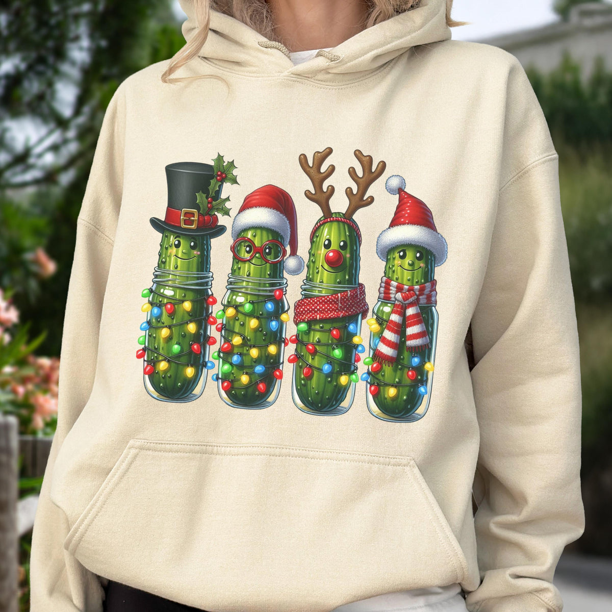 May include: A cream-colored hooded sweatshirt featuring a graphic print of four green pickles in glass jars. Each pickle is decorated with colorful string lights and festive holiday accessories including a top hat, Santa hat, and reindeer antlers.